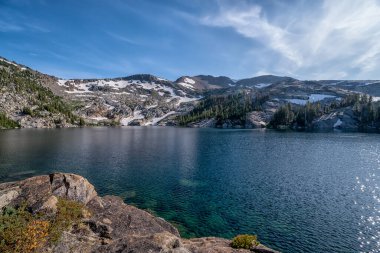 Arkasında dağ olan güzel bir göl. Su sakin ve berrak. Gökyüzü mavi ve bazı bulutlar var. Huzurlu ve huzurlu bir sahne.