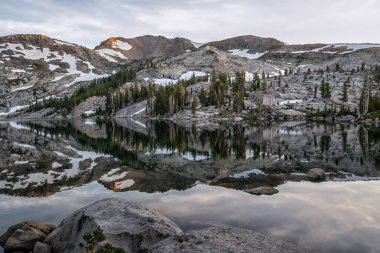 Önünde göl olan bir dağ sırası. Göl sakin ve uzaktaki dağları yansıtıyor. Gökyüzü bulutlu ve güneş batıyor, sakin ve huzurlu bir atmosfer yaratıyor.