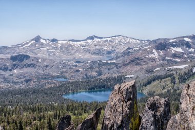 Önünde göl olan bir dağ sırası. Göl ağaçlar ve kayalarla çevrilidir. Gökyüzü açık ve mavi