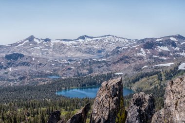 Önünde göl olan bir dağ sırası. Göl ağaçlar ve kayalarla çevrilidir. Gökyüzü açık ve mavi