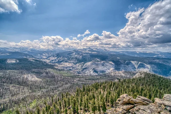 Arka planda bulutlu bir gökyüzü olan bir dağ sırası. Bulutlar beyaz ve kabarık, ve gökyüzü mavi. Ağaçlar yeşil ve yemyeşil ve dağlar karla kaplı. Huzur dolu bir sahne.