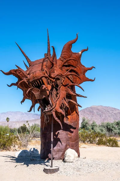 Çölde büyük, metalik bir ejderha heykeli oturuyor. Heykel kayalarla çevrili ve o bir sanat eseri. Çöl manzarası ve ejderhanın metalik görünüşü bir gizem hissi yaratıyor.
