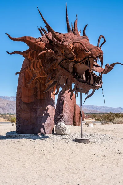 Çölde büyük, metalik bir ejderha heykeli duruyor. Ejderhanın tehditkar bir görünüşü var ve etrafı kayalarla çevrili. Sahne sessiz ve sakin.