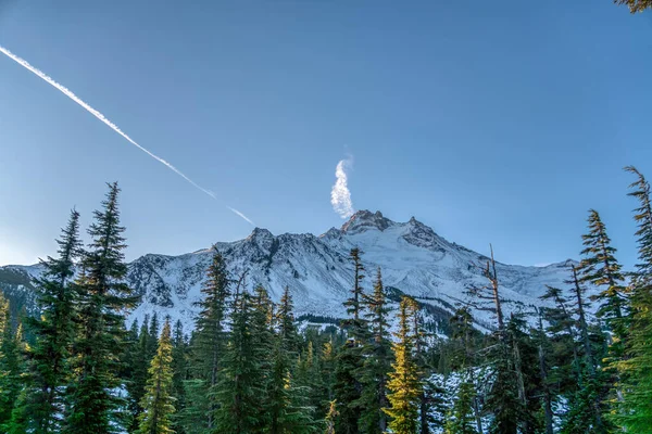 Arkasında açık mavi gökyüzü olan kar ve ağaçlarla kaplı bir dağ sırası. Gökyüzü bulutlarla dolu ve bir uçak uzaklarda uçuyor.