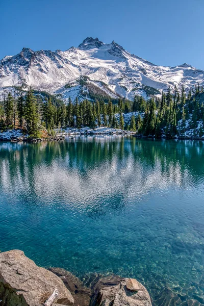 Bir dağ sırası bir göle yansıyor. Su berrak ve sakin. Gökyüzü mavi ve hiç bulut yok.