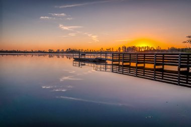 Arka planda günbatımı olan bir rıhtım var. Su sakin ve gökyüzü turuncu.