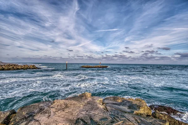 Okyanus sakin ve gökyüzü bulutlu. Su mavi ve kayalar gri.