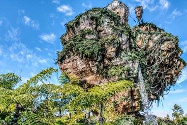 Üzerinde şelale olan büyük bir kaya. Kaya yeşil bitkilerle kaplı ve gökyüzü mavi