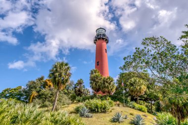 Arka planda palmiye ağaçları olan bir tepede kırmızı bir deniz feneri var. Sahne huzurlu ve sakin, deniz feneri mavi gökyüzüne karşı gururla ayakta duruyor.