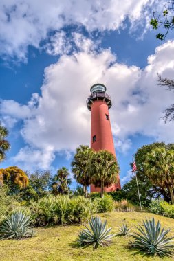 Yeşil çimenli bir alanı olan bir tepede kırmızı bir deniz feneri var. Gökyüzü bulutlu ve güneş parlıyor