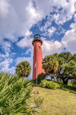Kırmızı bir deniz feneri, palmiye ağaçları ve otlarla dolu bir tepede. Gökyüzü mavi ve bulutlu.