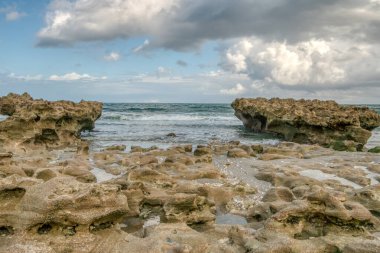 Arka planda büyük bir su kütlesi olan kayalık bir sahil. Kayalar sahile dağılmış, engebeli ve doğal bir manzara yaratıyorlar. Gökyüzü bulutlu, derinlik katıyor