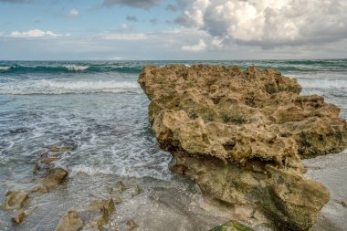 Önünde büyük bir kaya olan kayalık bir sahil. Gökyüzü bulutlu ve su dalgalı.