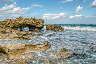 Okyanus sakin ve kayalar sudan dışarı fırlıyor. Gökyüzü mavi ve bazı bulutlar var.