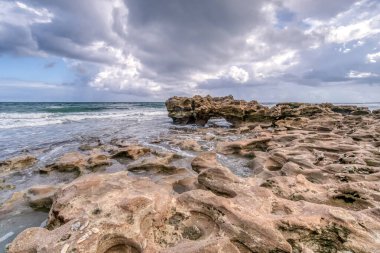 Arka planda bulutlu bir gökyüzü olan kayalık bir sahil. Kumsal kayalık ve içinde bir sürü delik var.