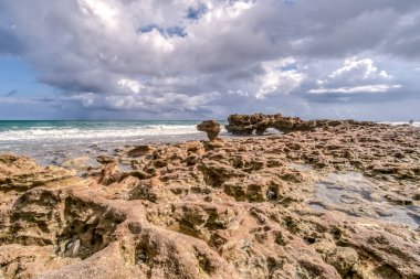 Arka planda bulutlu bir gökyüzü olan kayalık bir sahil. Kayalar sahilin her tarafına dağılmış ve dalgalar kıyıya çarpıyor.