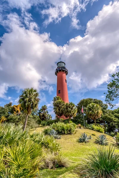 Arka planda palmiye ağaçları olan bir tepede kırmızı bir deniz feneri var. Gökyüzü bulutlu, ama güneş bulutların arasından parlıyor. Sahne huzurlu ve sakin, deniz feneri dimdik ayakta.