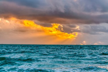 Gökyüzü bulutlu ve güneş batıyor. Güneş bulutların arasından parlıyor, güzel bir turuncu ve pembe gökyüzü yaratıyor. Okyanus sakin ve su mavi.