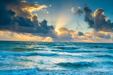 Gökyüzü bulutlu ve güneş bulutların arasında parlıyor. Okyanus sakin ve su mavi.