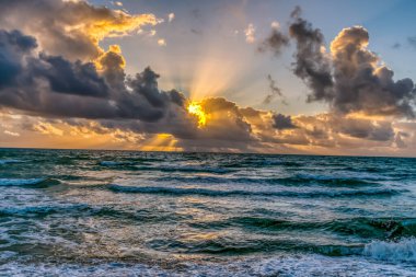 Gökyüzü bulutlu ve güneş batıyor. Okyanus sakin ve su koyu mavi bir renktir.
