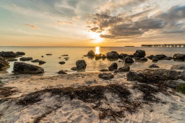 Büyük bir kaya tarlası ve küçük bir iskelesi olan bir sahil. Güneş batıyor ve gökyüzü bulutlu.