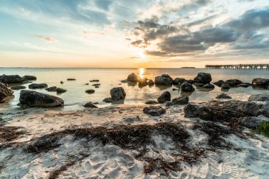 Büyük bir kaya tarlası ve küçük bir iskelesi olan bir sahil. Gökyüzü bulutlu ve güneş batıyor.