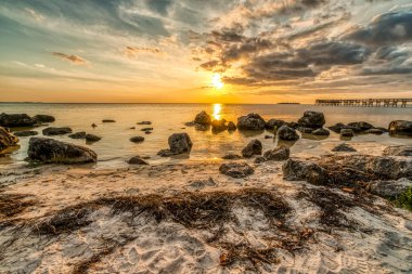 Arkasında büyük bir kaya tarlası ve iskelesi olan bir sahil. Gökyüzü bulutlu ve güneş batıyor.