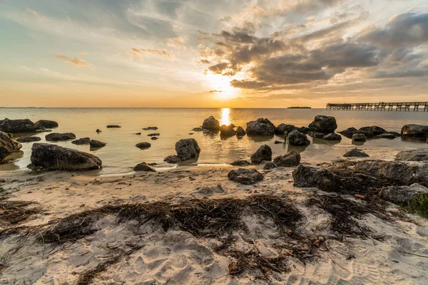 Büyük bir kaya tarlası ve küçük bir iskelesi olan bir sahil. Güneş batıyor ve gökyüzü bulutlu.