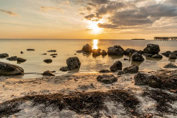 Arka planda büyük bir su kütlesi olan kayalık bir sahil. Güneş batıyor, sahne üzerine sıcak bir parıltı saçıyor.