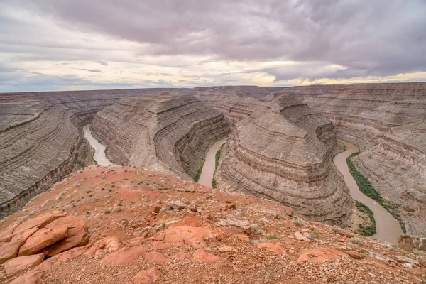 Kayalık, kırmızı bir manzarayla bir kanyonun içinden akan bir nehir. Gökyüzü bulutlu, karamsar bir atmosfer yaratıyor. Nehir kanyonda süzülüyor.