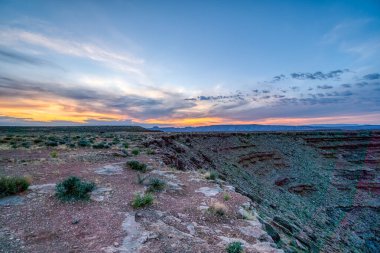 Çöl manzarasında güzel bir gün batımı. Gökyüzü bulutlarla dolu ve güneş batıyor, karaya sıcak bir parıltı saçıyor. Huzurlu ve huzurlu bir sahne.