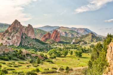 Ortasında büyük kırmızı bir kaya oluşumu olan bir dağ sırası. Dağlar ağaçlarla kaplı ve çimenler yemyeşil. Gökyüzü açık ve güneş parlıyor.