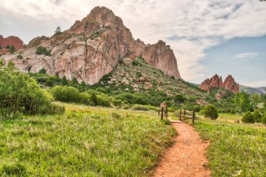 Arka planda dağ olan çimenli bir tarladan geçen bir patika. Gökyüzü bulutlu, ama güneş hala parlıyor.