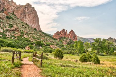 Sol tarafında çit olan çimenli bir araziye giden bir patika var. Gökyüzü bulutlu ve arkaplandaki dağlar yeşil çimenlerin güzel bir kontrastı.