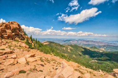 Açık mavi gökyüzü ve birkaç bulutu olan bir dağ sırası. Gökyüzü çok parlak ve dağlar ağaçlarla kaplı.