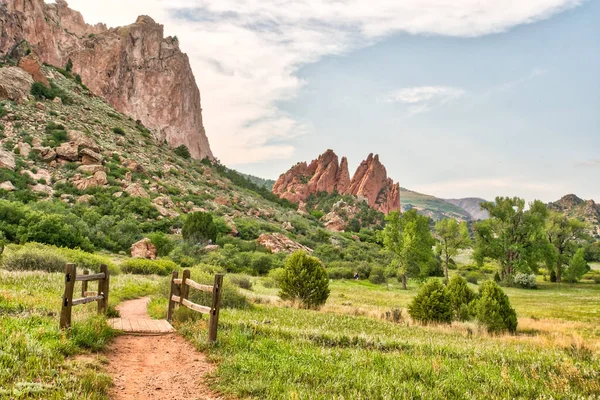 Sol tarafında çit olan çimenli bir araziye giden bir patika var. Gökyüzü bulutlu ve arkaplandaki dağlar yeşil çimenlerin güzel bir kontrastı.