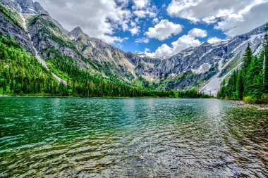 Dağlarla çevrili güzel bir göl. Su sakin ve berrak, yukarıdaki mavi gökyüzünü yansıtıyor. Sahne sakin ve huzurlu, arka planda dağlar ihtişam hissi katıyor.