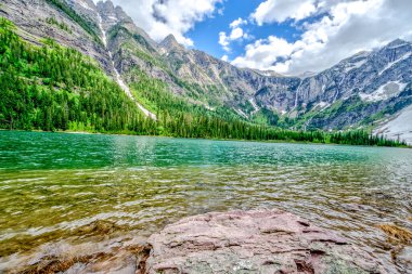 Arkasında dağlar olan güzel bir göl. Su berrak ve sakin. Dağlar ağaçlarla kaplı ve gökyüzü mavi. Huzurlu ve huzurlu bir sahne.