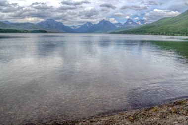 Arkasında dağlar olan bir göl. Su sakin ve berrak. Gökyüzü bulutlu ve dağlar uzakta.