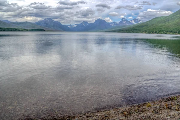 Arkasında dağlar olan bir göl. Su sakin ve berrak. Gökyüzü bulutlu ve dağlar uzakta.