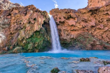 Bir şelale mavi bir su havuzuna akıyor. Su berrak ve sakin ve havuzun etrafındaki kayalar pürüzlü ve pürüzsüz. Sahne huzurlu ve huzurlu.