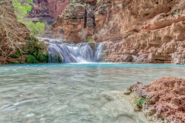Güzel bir şelale açık mavi bir havuza akıyor. Su sakin ve havuzun etrafındaki kayalar kahverengidir. Huzurlu ve huzurlu bir sahne.