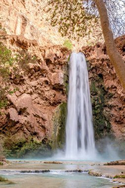 Arka planda yemyeşil bir orman bulunan bir şelale nehre akıyor. Su berrak ve şelaleden gelen sis dingin bir atmosfer yaratıyor.