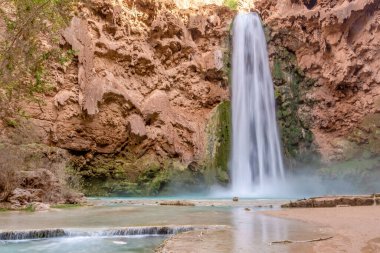 Bir şelale su havuzuna akıyor. Su temiz ve havuzun etrafındaki kayalar kahverengi.