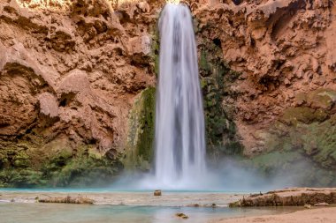 Bir şelale su havuzuna akıyor. Su temiz ve havuzun etrafındaki kayalar kahverengi.