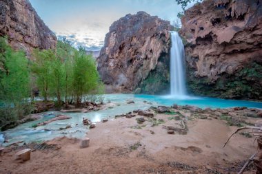 Güzel bir şelale yemyeşil bir ormanla çevrilidir. Su kristal gibi berrak ve kayalar etrafa saçılmış durumda. Sahne huzurlu ve huzurlu.