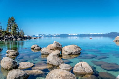 Arka planda bir su kütlesi olan kayalık bir kıyı şeridi. Su sakin ve berrak ve kayalar tüm bölgeye dağılmış durumda. Gökyüzü masmavi ve huzur dolu.