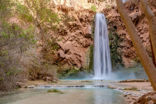 Arka planda bir ağaç olan bir şelale göle akıyor. Su berrak ve sakin. Çevresi yemyeşil ve yemyeşil. Huzurlu ve huzurlu bir sahne.