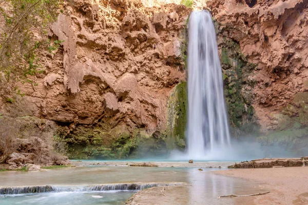Bir şelale su havuzuna akıyor. Su temiz ve havuzun etrafındaki kayalar kahverengi.