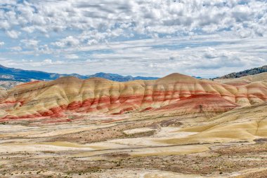 Kırmızı, sarı ve kahverengi renk desenli bir dağ sırası. Gökyüzü bulutlu ve güneş parlıyor